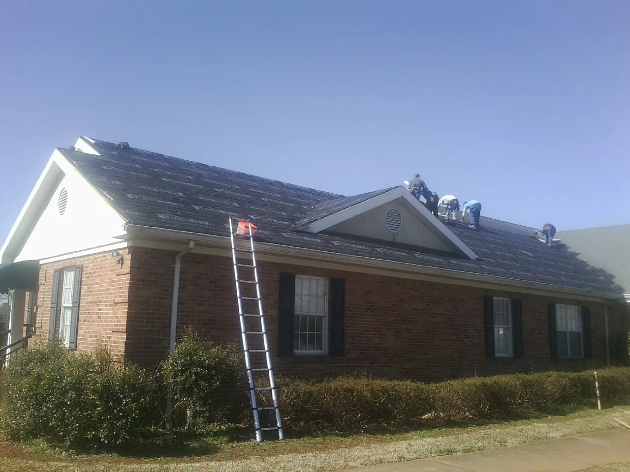 Crew tearing shingles off a brick ranch roof with a ladder set — Emergency Roof Repair work by our crew in Broomfield