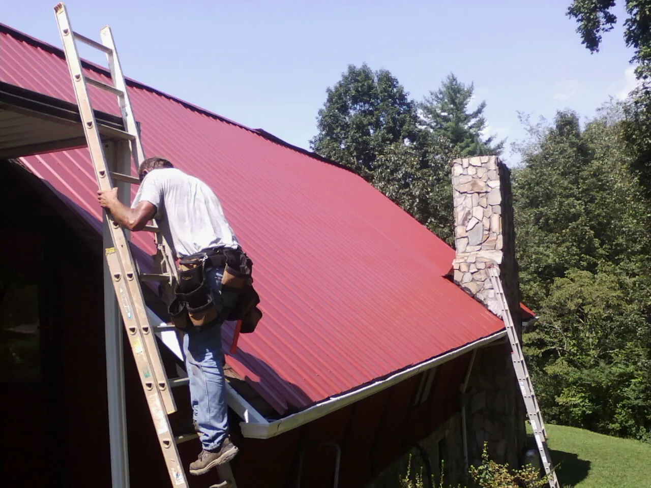 Roofer on a ladder beside a red metal roof and stone chimney — Soffit & Fascia Repair by Roofing Brothers in Broomfield